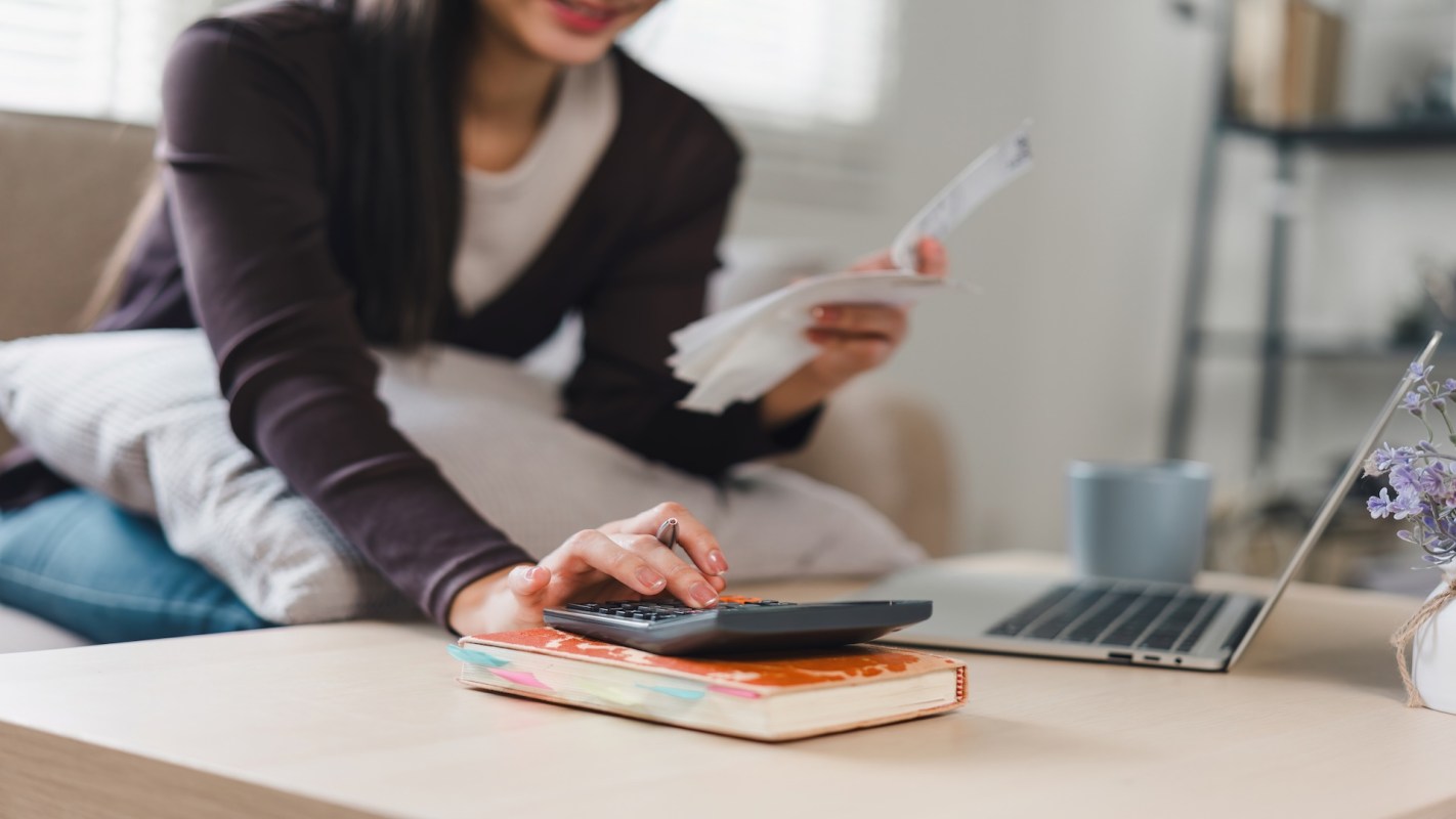 A woman uses a calculator while holding receipts, with a laptop and a cup nearby on a table.