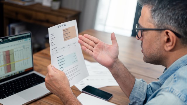 A man, sitting at a desk, expresses exasperation with a raised hand while reviewing a utility bill.