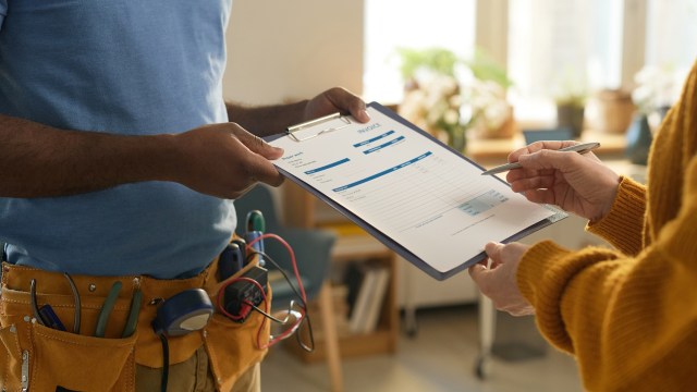 A person with a tool belt hands an invoice to another person in an indoor setting.