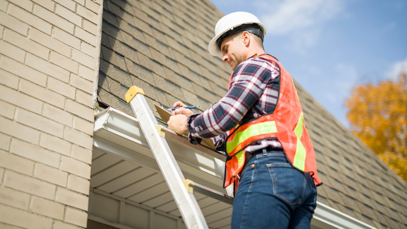 A worker in safety gear inspecting a roof and taking notes on a clipboard.