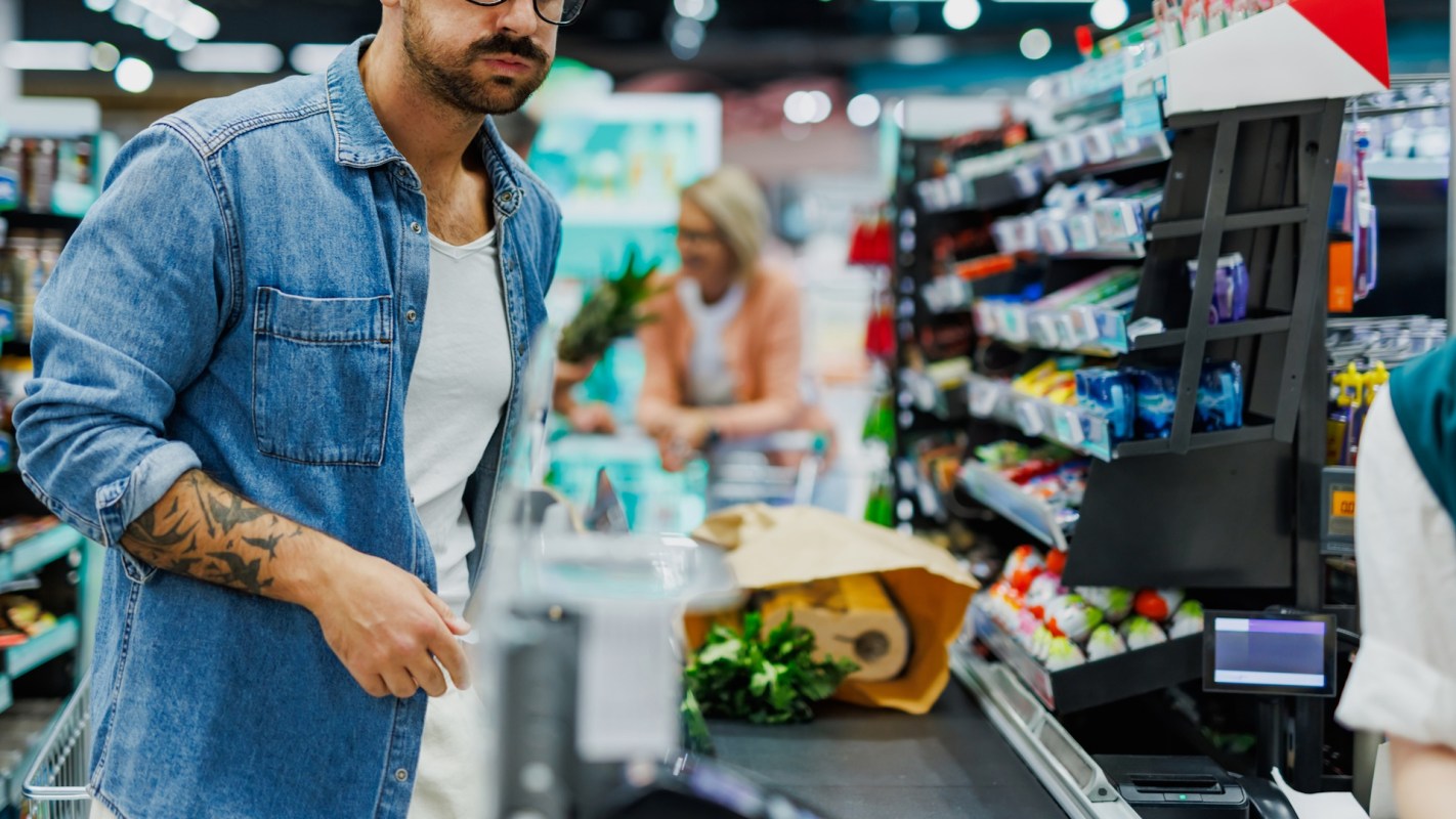 A man stands at a checkout counter in a grocery store with bags of groceries.
