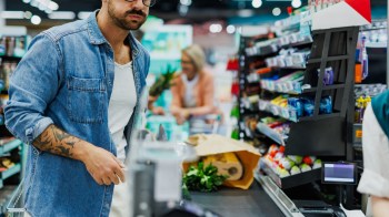 A man stands at a checkout counter in a grocery store with bags of groceries.