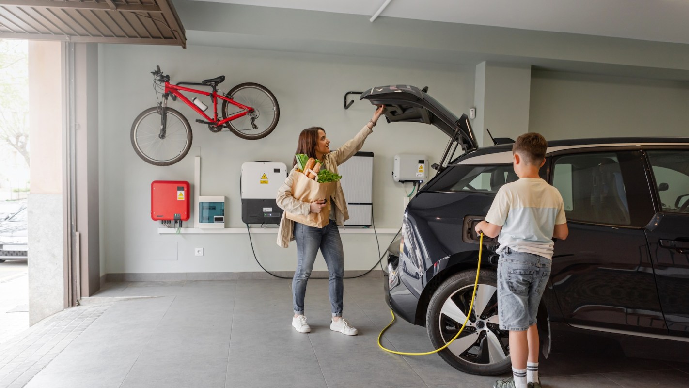 A woman in a garage places groceries in an electric vehicle while a boy connects a charging cable.