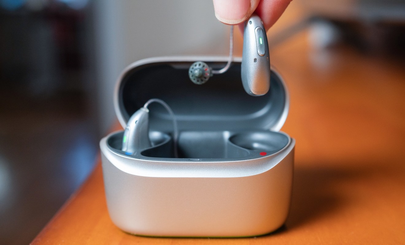 A person holds a hearing aid above an open charging case on a wooden surface.