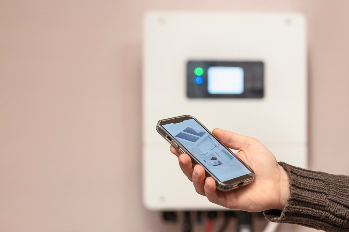 A hand holding a smartphone displaying an image in front of a mounted device on a wall.