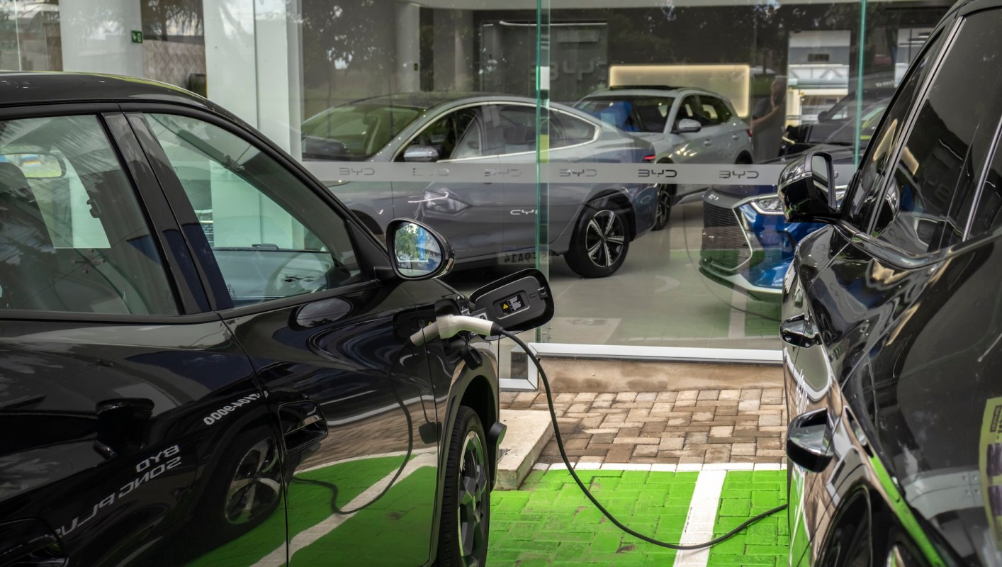 A black electric car charges at a station with other new vehicles visible in the background of a dealership.