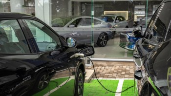 A black electric car charges at a station with other new vehicles visible in the background of a dealership.