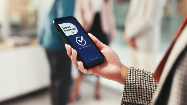 A person holds a smartphone displaying a travel insurance app in a busy setting.