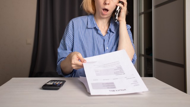 A woman is on the phone while holding a utility bill and looking surprised at a table with a calculator.