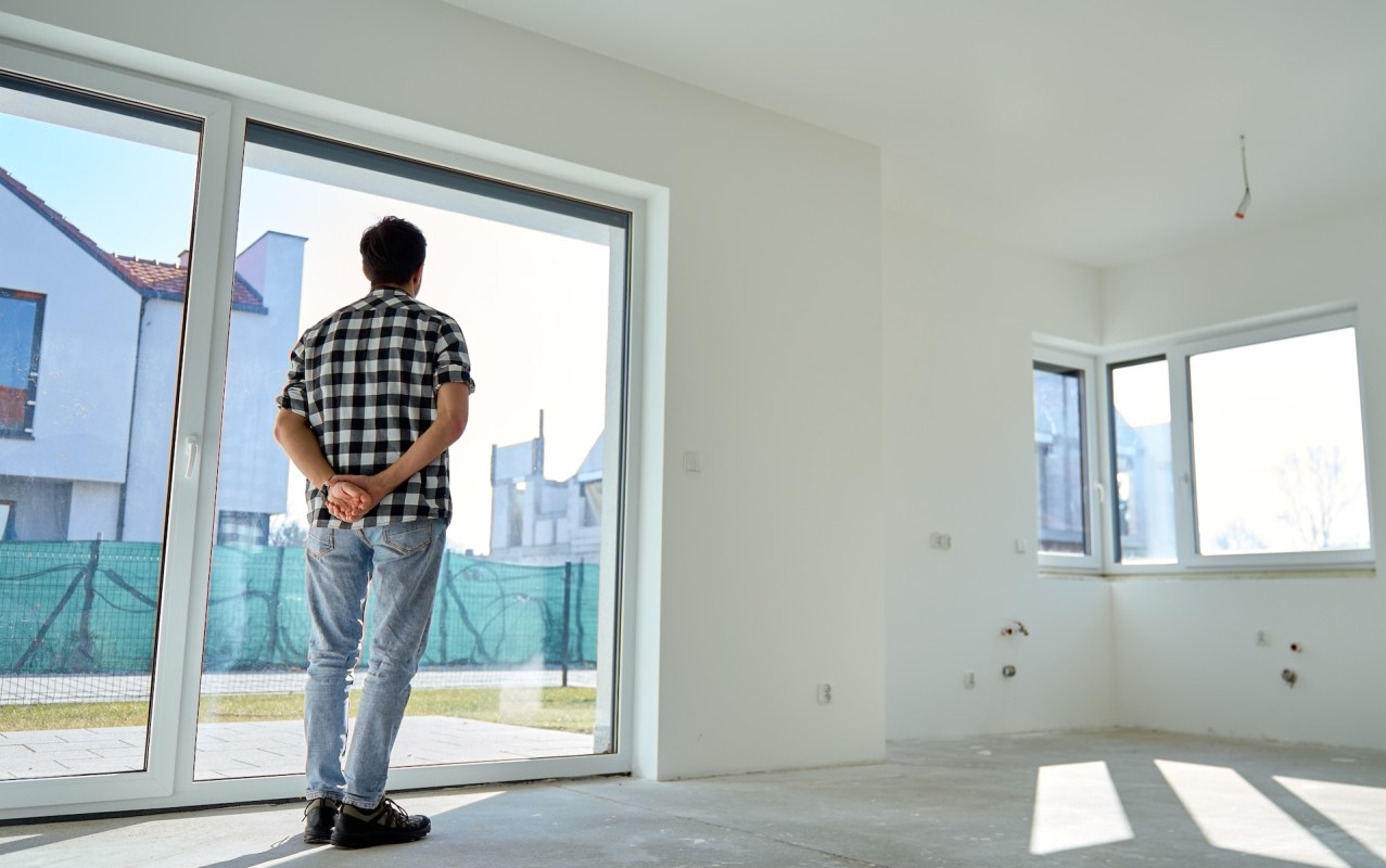 A person stands inside an empty unfinished room, looking out through large windows at a construction site.