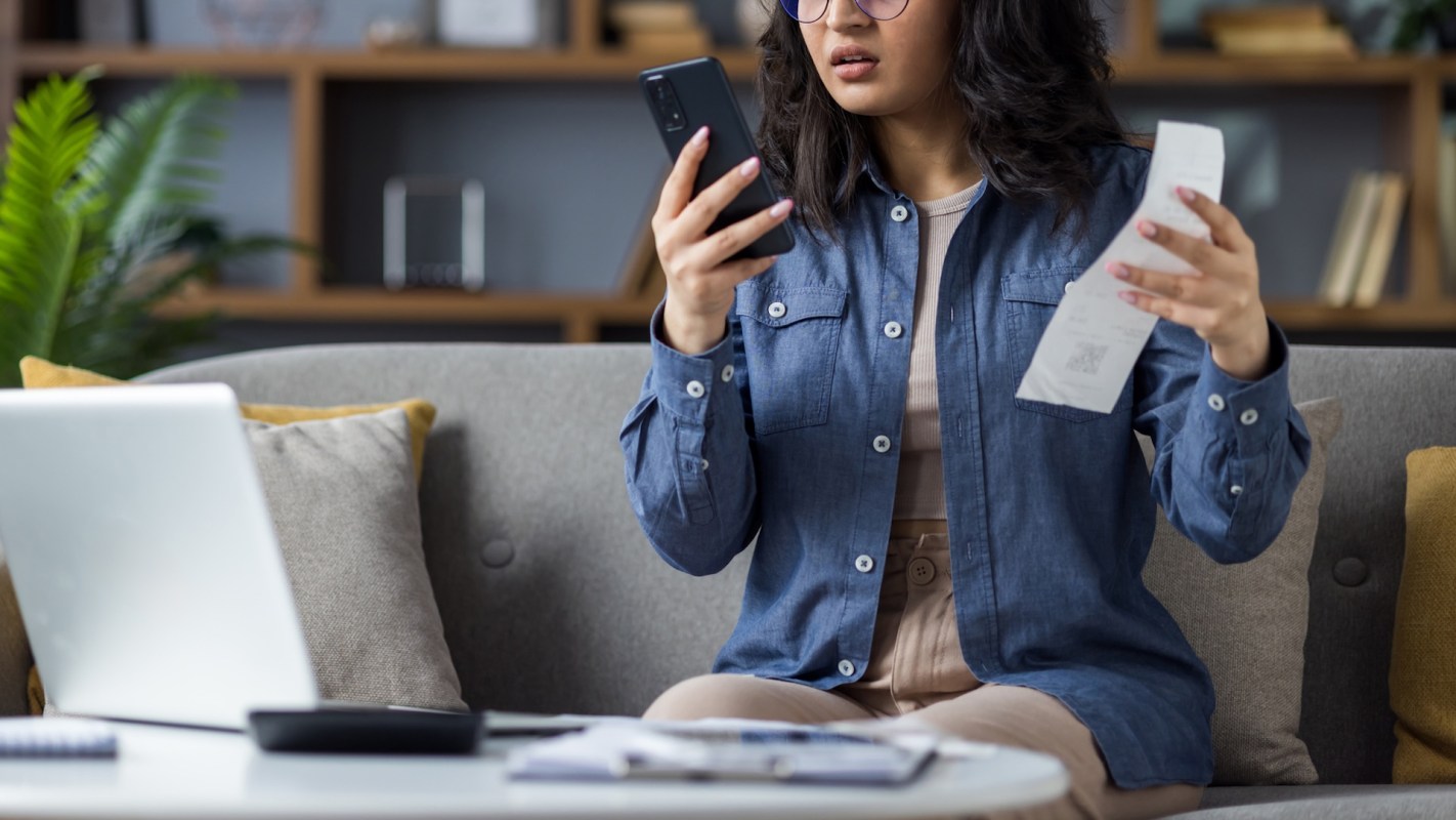 A woman looks annoyed while looking at her phone and holding a receipt.