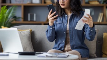 A woman looks annoyed while looking at her phone and holding a receipt.
