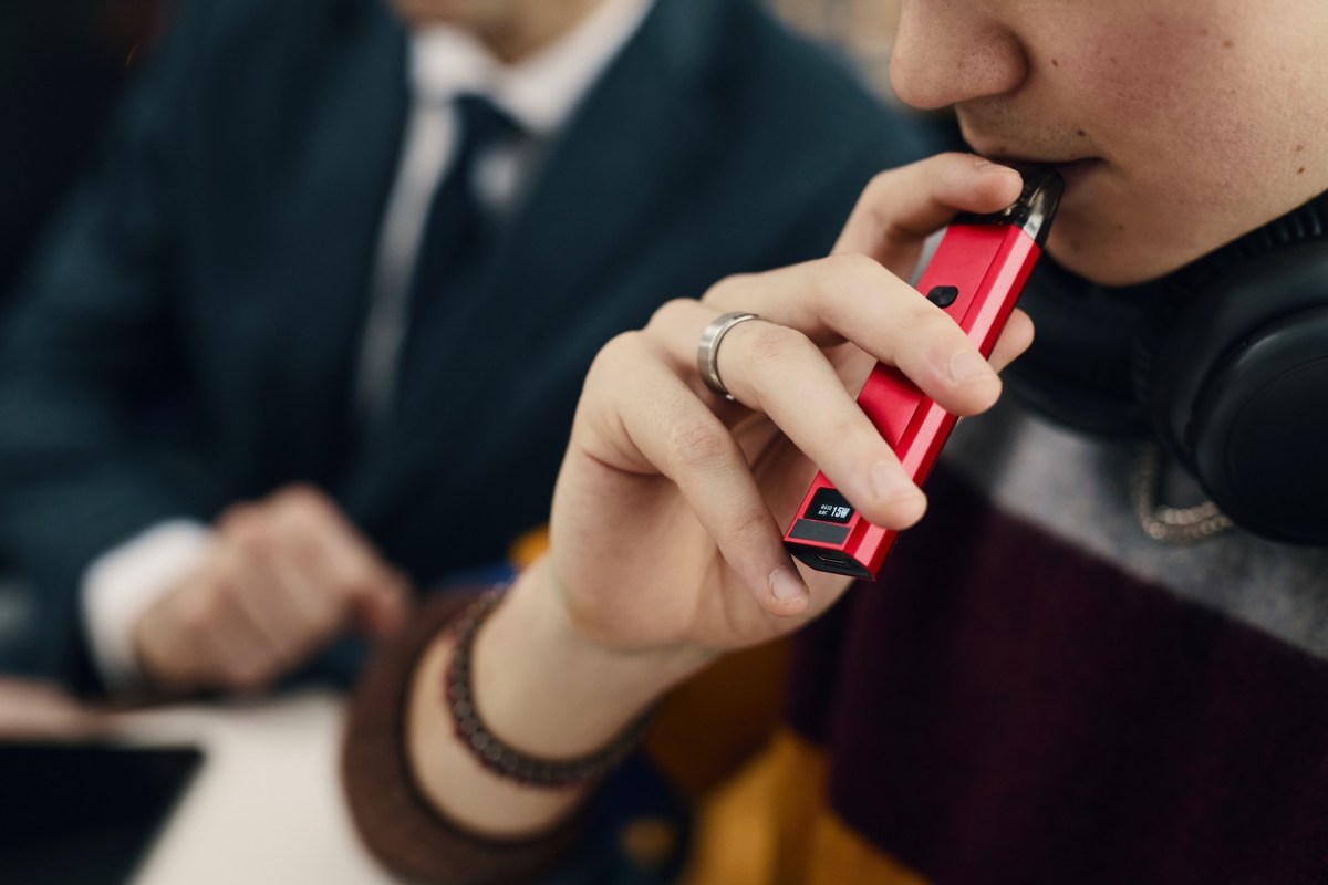 A person holds a red vape pen while sitting at a table with another individual in the background.
