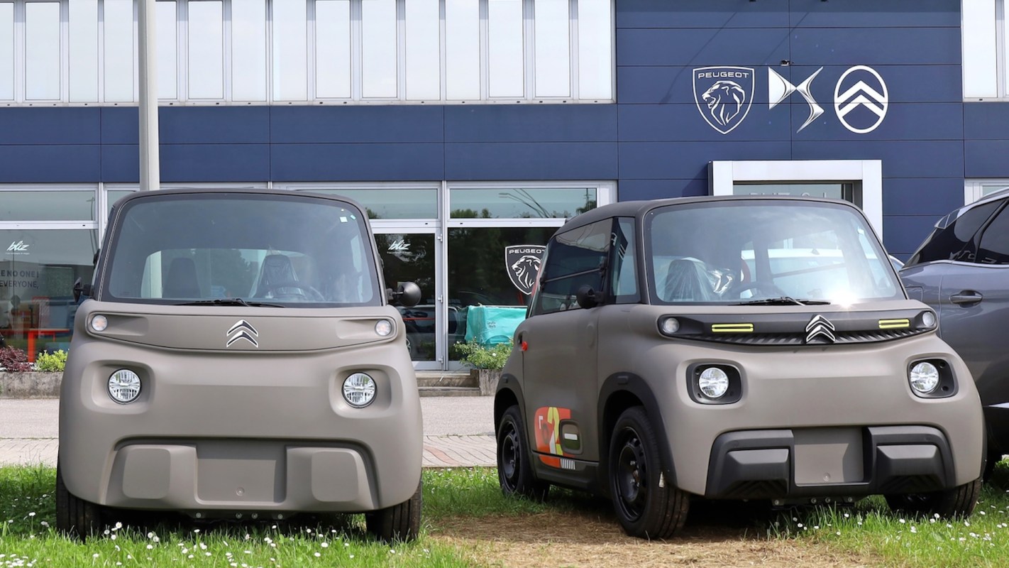 Two compact electric cars parked on grass outside a building with Peugeot automotive branding.