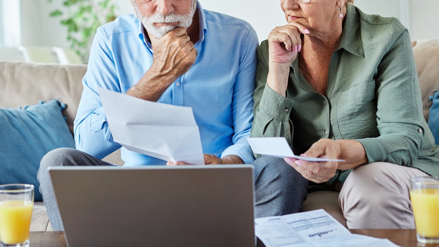 An elderly couple reviews documents together while seated on a couch beside a laptop.