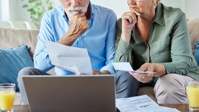 An elderly couple reviews documents together while seated on a couch beside a laptop.
