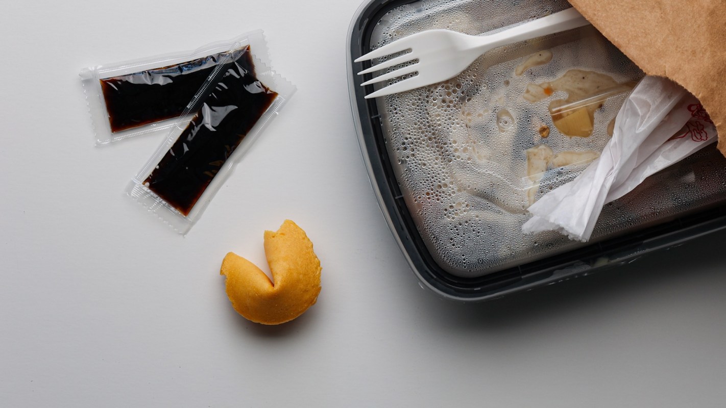 A takeout container with remnants of food, two packets of sauce, and a broken fortune cookie.