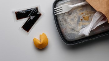 A takeout container with remnants of food, two packets of sauce, and a broken fortune cookie.