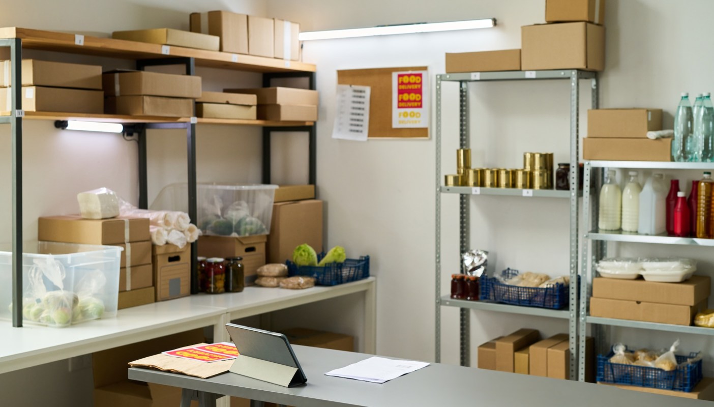 A storage room with shelves containing boxes, jars, and various food items, plus a tablet on a table.