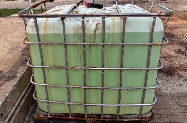 An intermediate bulk container with green liquid, surrounded by rusted metal and concrete flooring.
