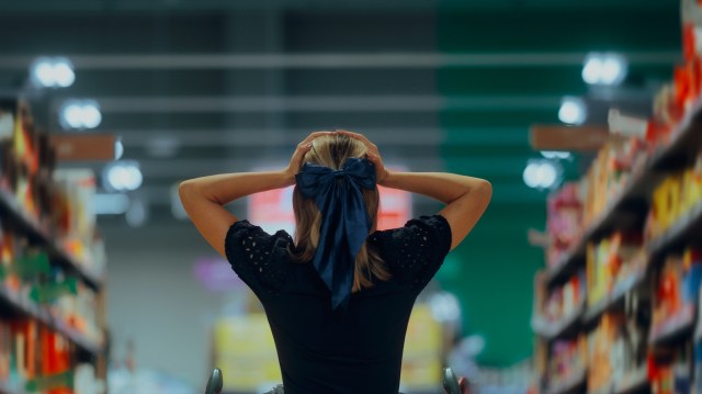 A woman stands in a supermarket aisle with her hands on her head.