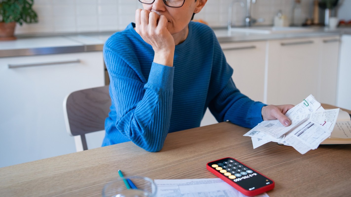A person at a kitchen table with receipts and a calculator, hand under chin, looking worried.