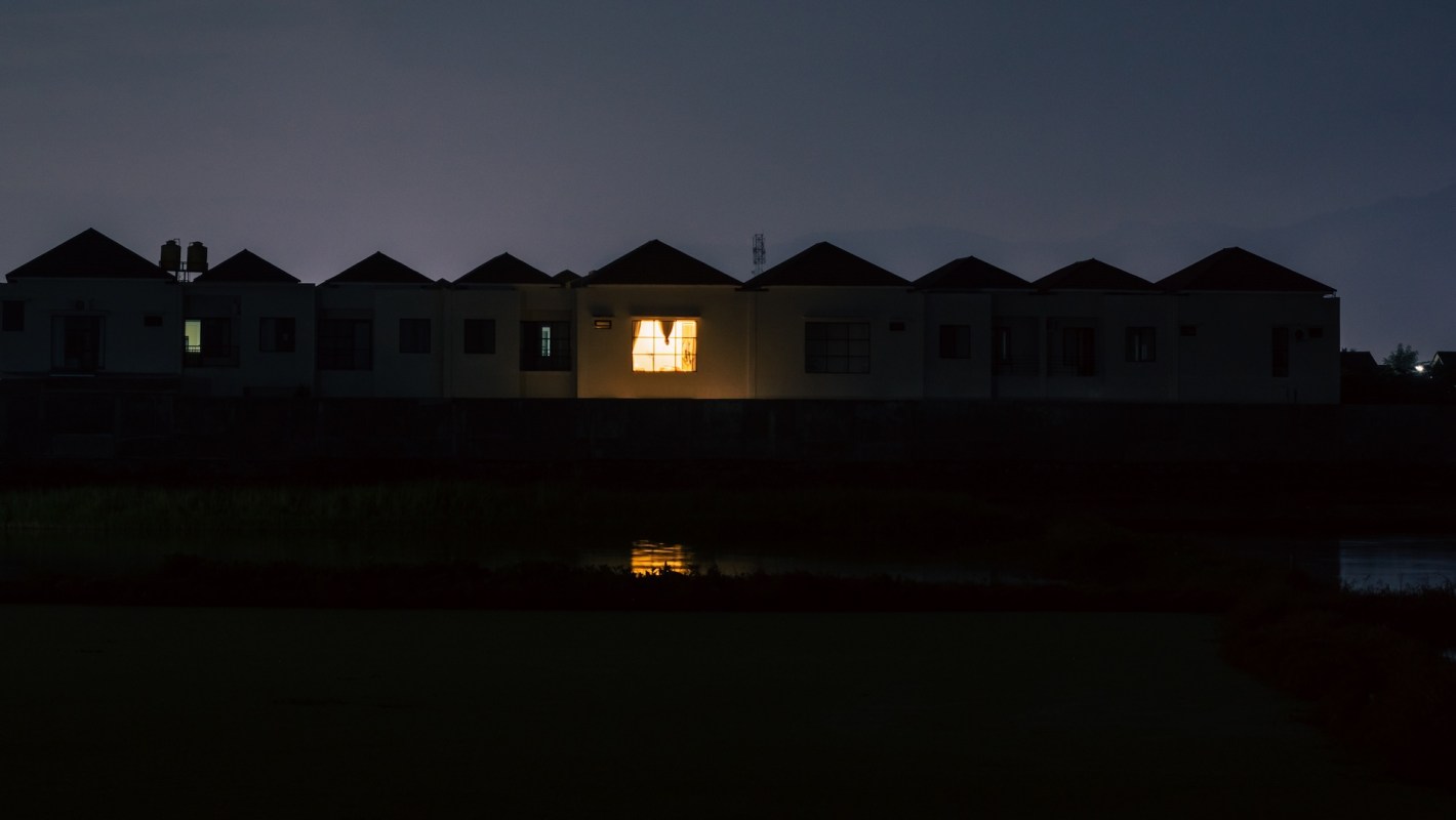 A row of houses at night with one illuminated window reflecting on the water.