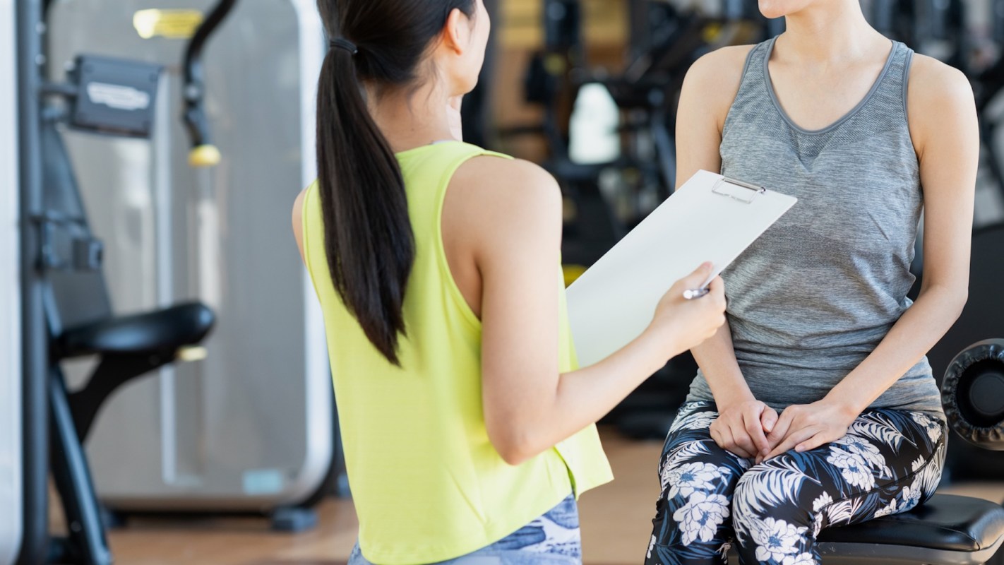 A trainer holds a clipboard while speaking with a client in a gym setting.