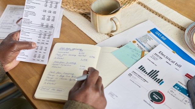 A person reviews financial documents as well as gas and electricity bills on a table with a cup beside them.