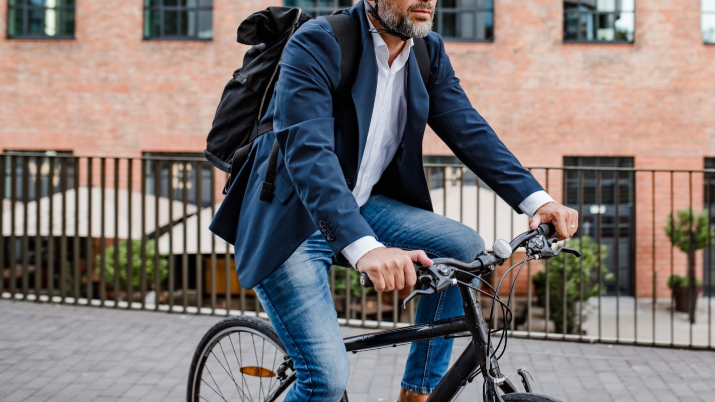 A person in a suit rides a bicycle on a paved path near a brick building.