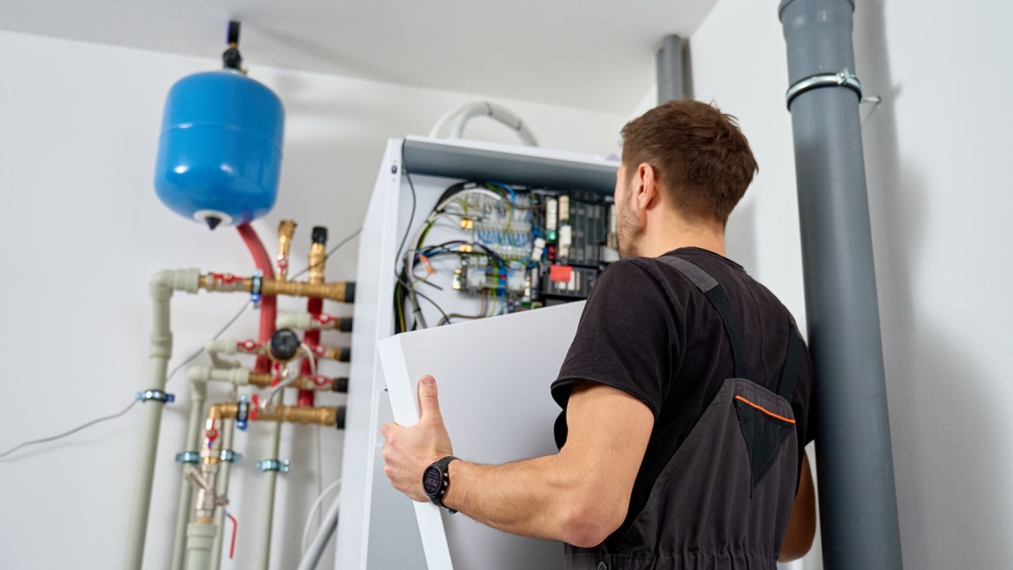 A person installs a heat pump in a utility room with pipes and a pressure tank.