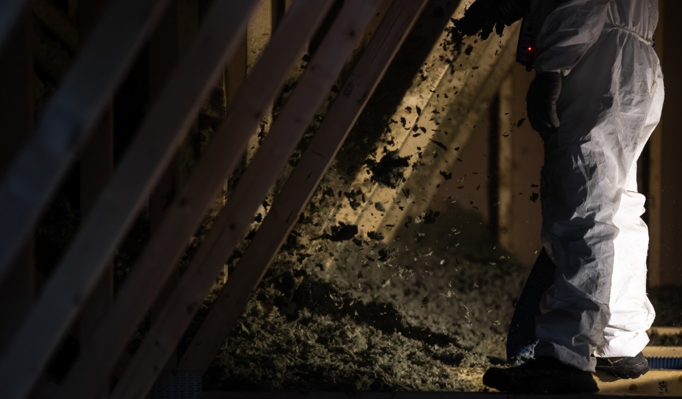A person in protective clothing works in an attic, adding insulation material.