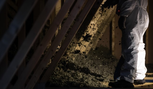 A person in protective clothing works in an attic, adding insulation material.
