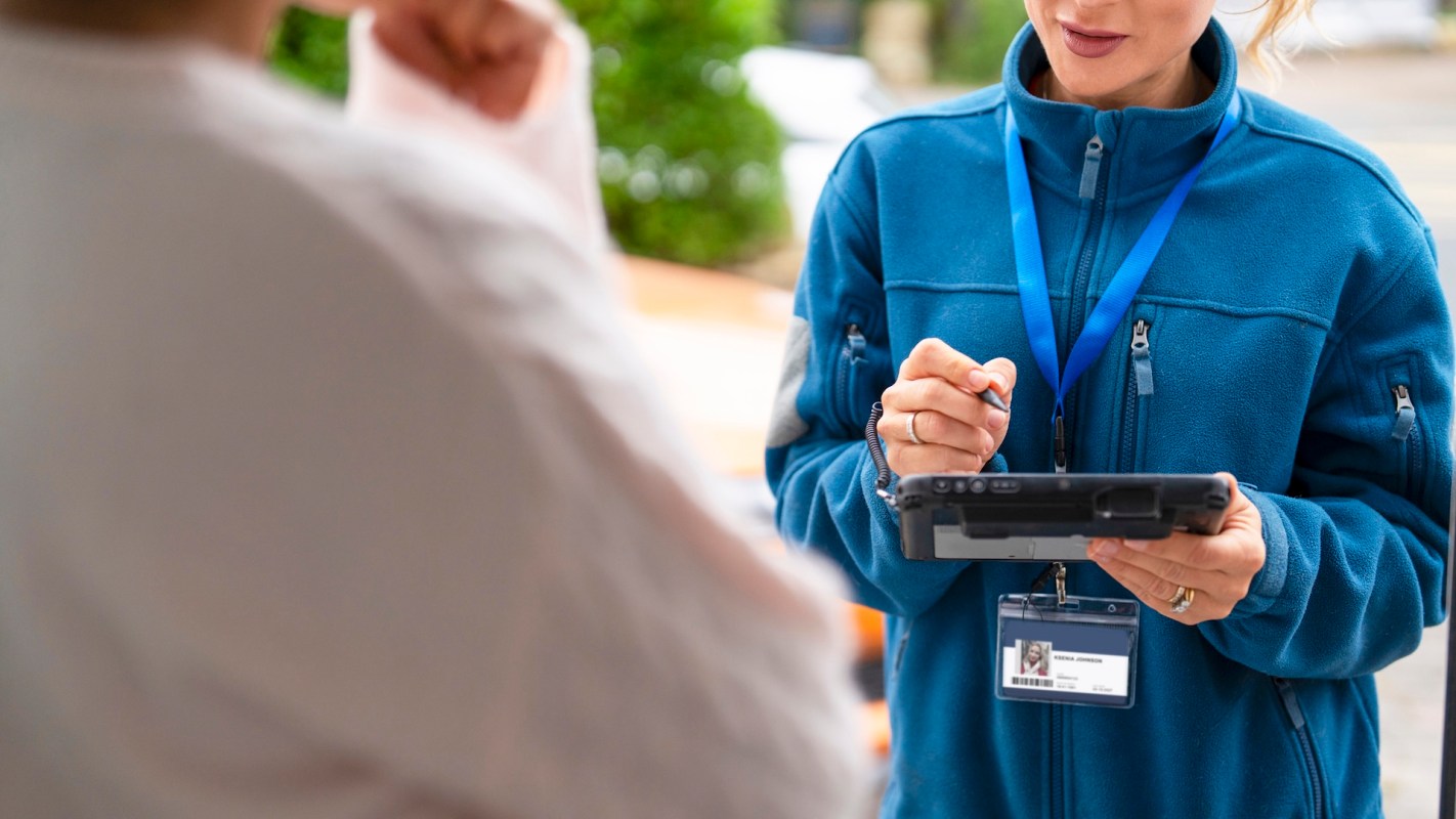 A person in a blue jacket at a door uses a tablet while engaging with another individual.