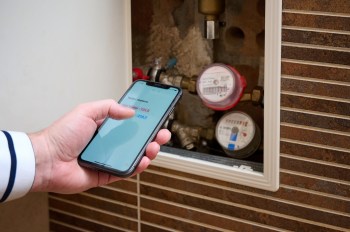A person uses a smartphone to check water meter readings beside visible plumbing and water meters.