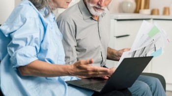A senior woman and a man discuss documents while looking at a laptop.