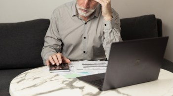 An older man, with a hand on his head, uses a calculator while working on documents at a table with a laptop nearby.