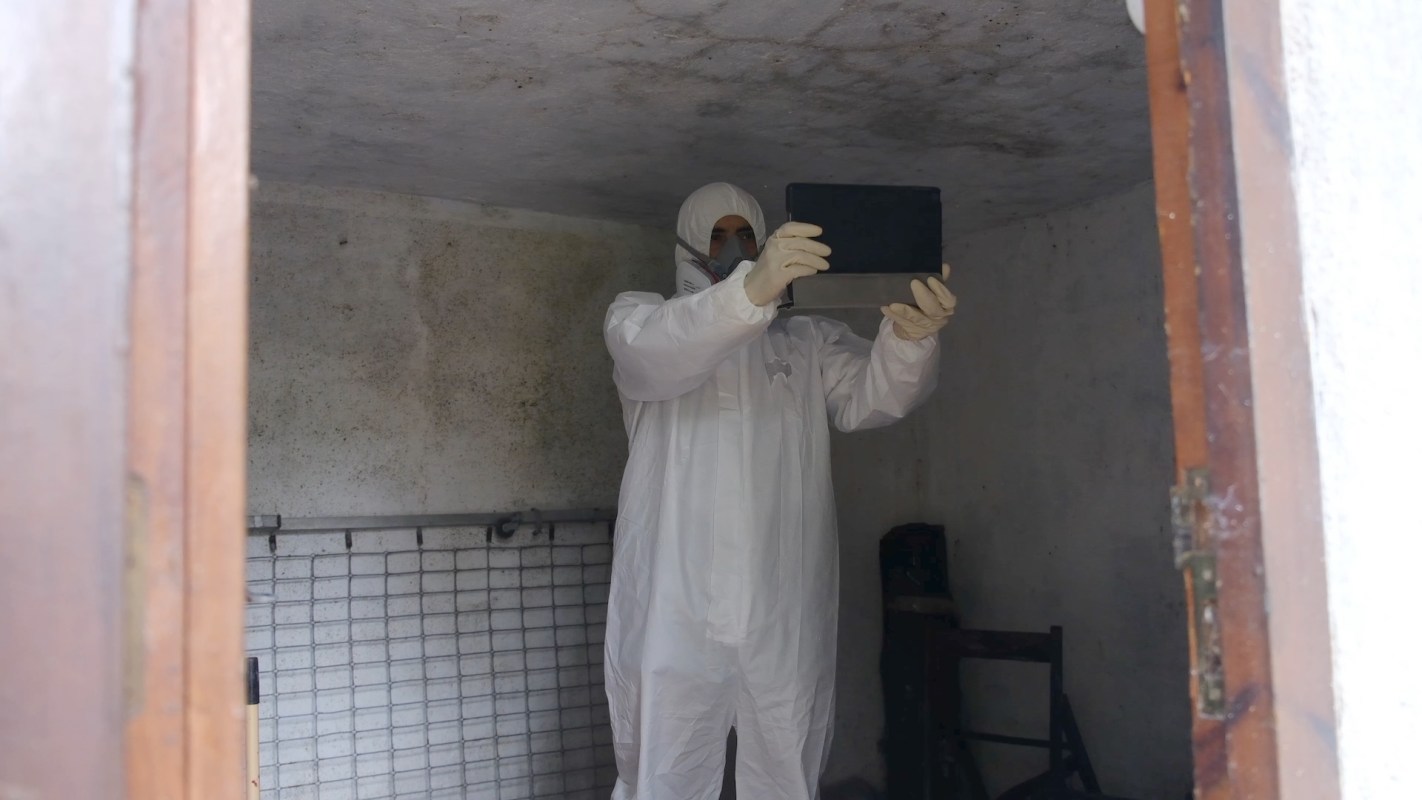 A person in protective clothing holds a tablet inside a bare room.