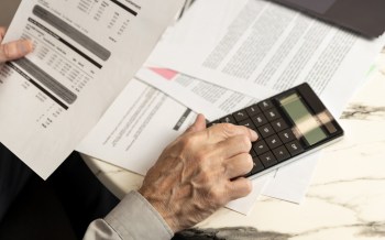 An elderly man uses a calculator while holding financial documents on a table.