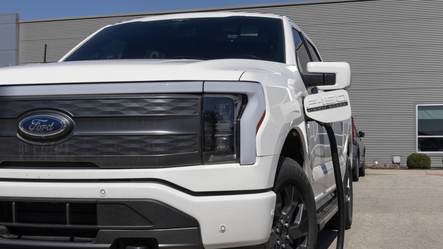 A white Ford F-150 Lightning parked outside a building, with a charging cord plugged into it.