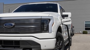 A white Ford F-150 Lightning parked outside a building, with a charging cord plugged into it.