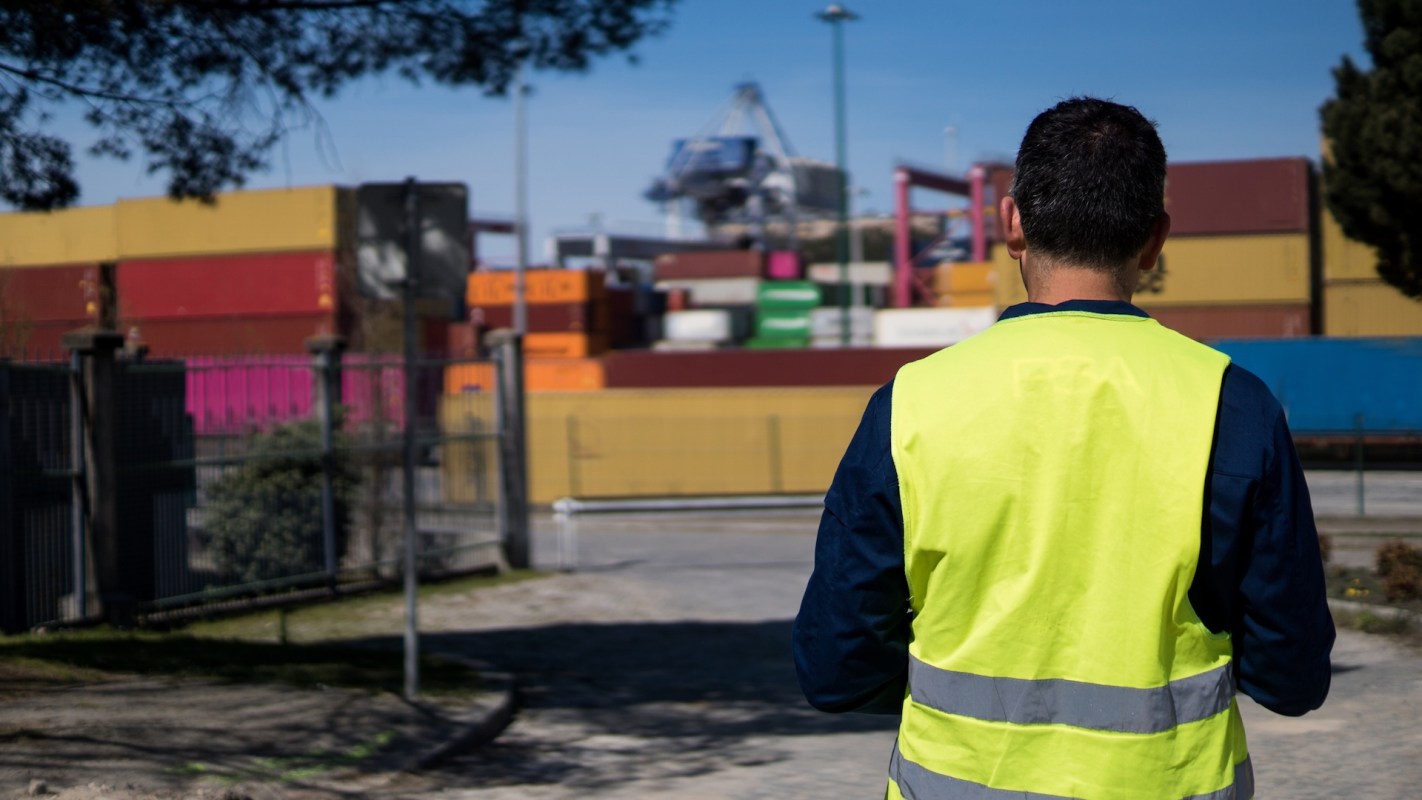 A person in a high-visibility vest stands in front of shipping containers at a port.