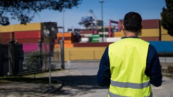 A person in a high-visibility vest stands in front of shipping containers at a port.