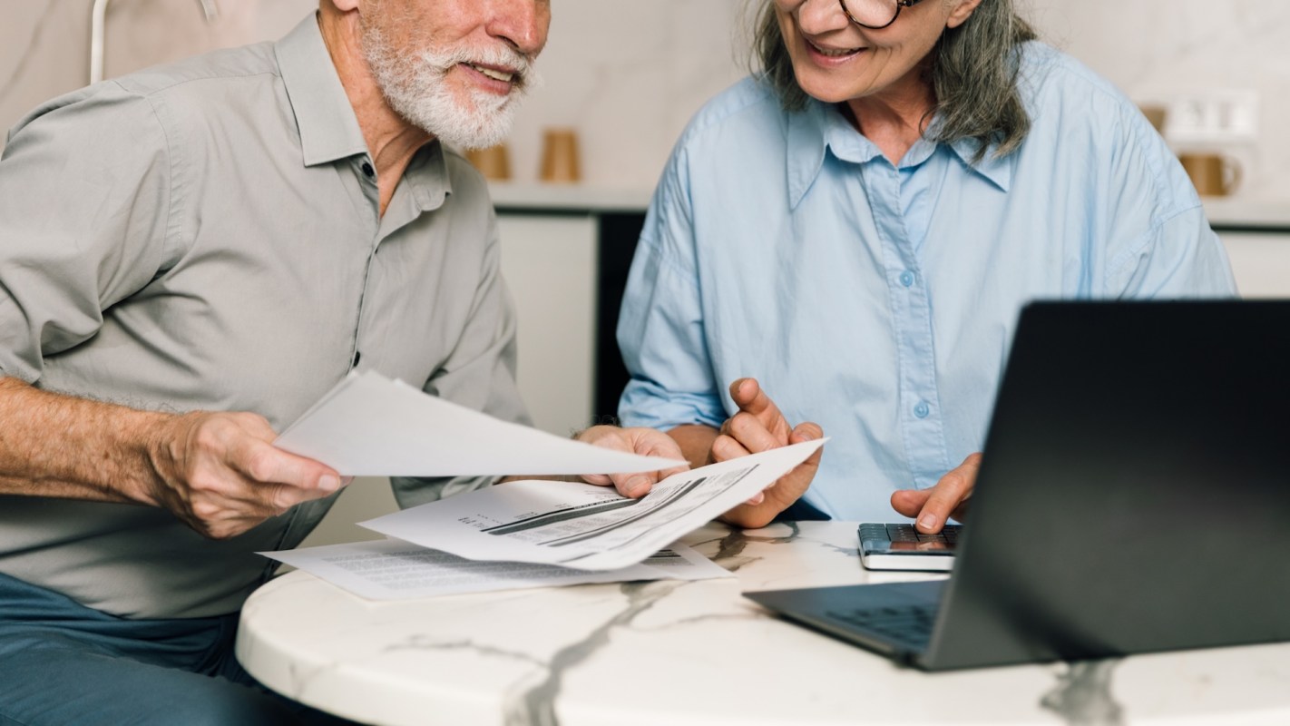 An elderly couple smiles while reviewing documents together at a table with a laptop and calculator.