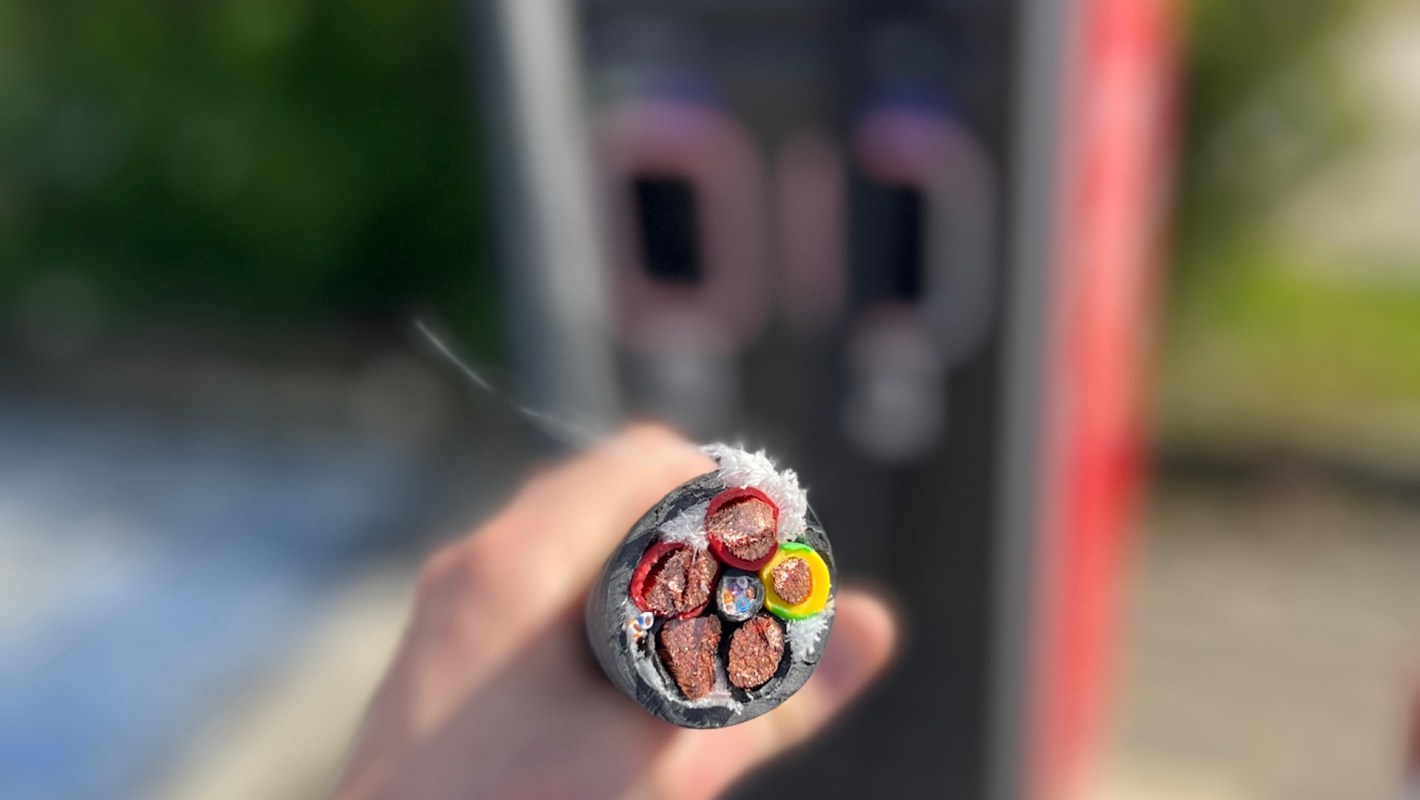 Outside of an EV charger, a hand holds a section of electrical cable with multiple colored wires inside.