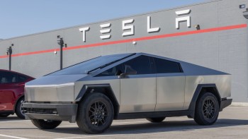 A Tesla Cybertruck parked next to a red Tesla vehicle in front of a gray building with the Tesla logo.