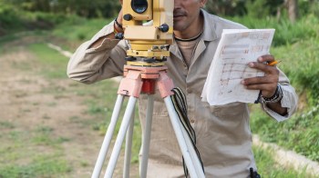 A man operates a surveying instrument while holding a set of construction plans outdoors.