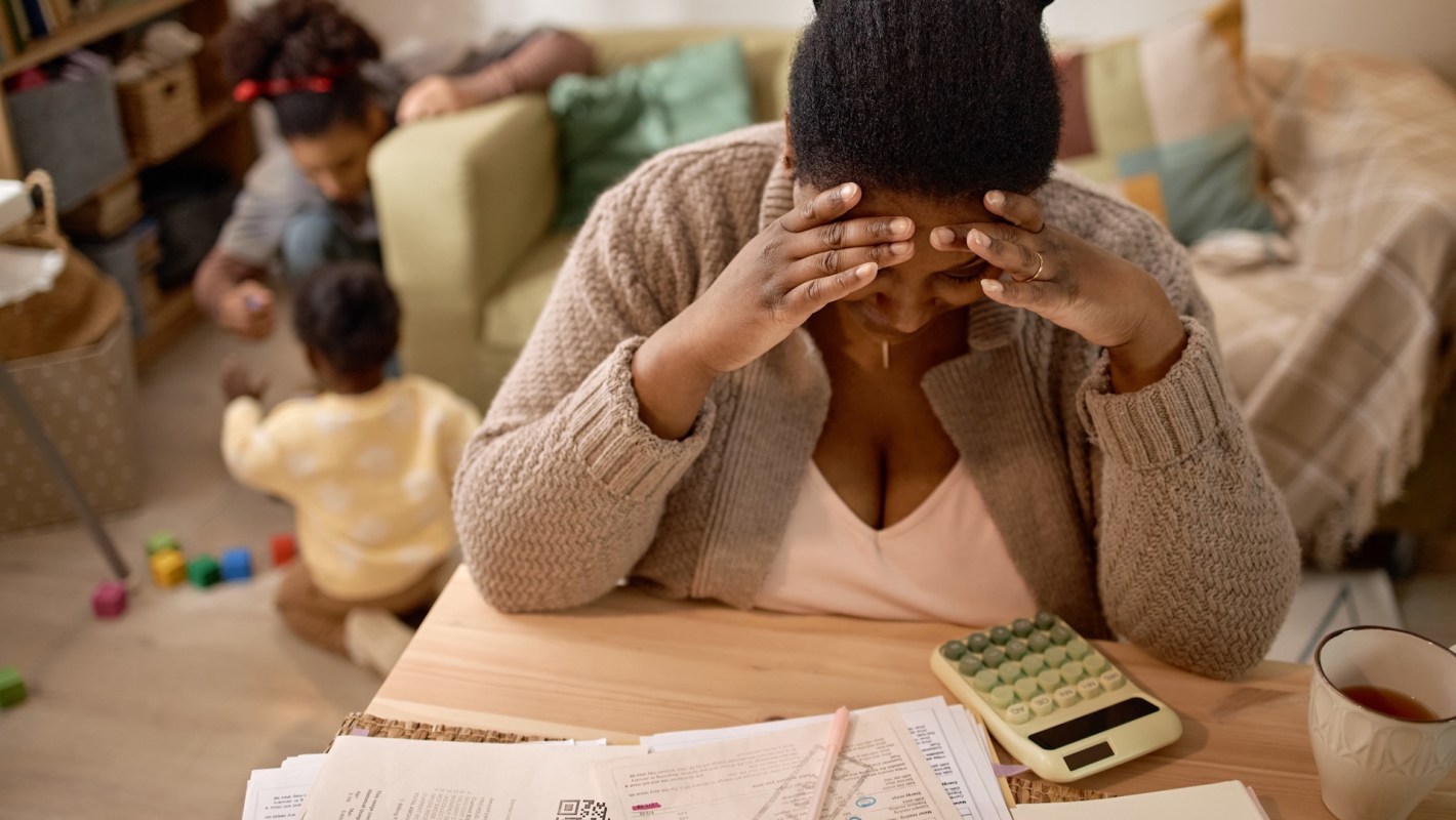 A woman sits at a table with papers and a calculator, her hands on her head, while two children play nearby.