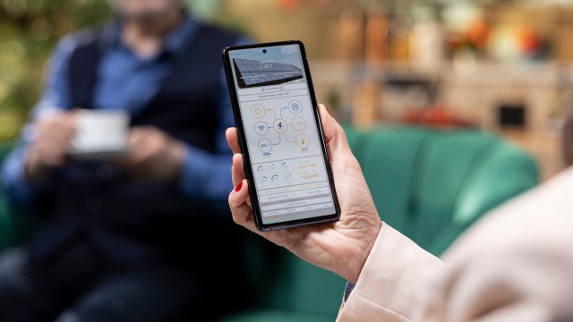 A person holds a smartphone displaying energy data while another person sits nearby with a cup.