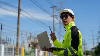 A construction worker holds a laptop and a radio near power lines under a clear blue sky.